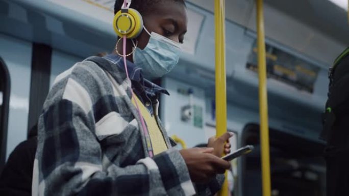 戴着医用口罩的年轻黑人妇女晚上乘地铁回家，用智能手机打字。时尚的女性乘坐公共交通工具旅行，尊重检疫规
