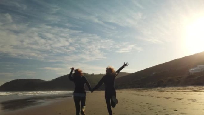 两个女性朋友的慢动作感到自由，在日落时分带着白色沙滩的荒岛海滩上，手拉手奔向蓝色的大海。