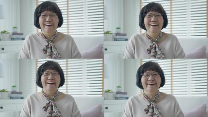 头部拍摄肖像快乐健康的中年亚洲妇女，戴眼镜坐在家里的沙发上。老太太笑着，微笑着看着客厅的摄像机。屏幕