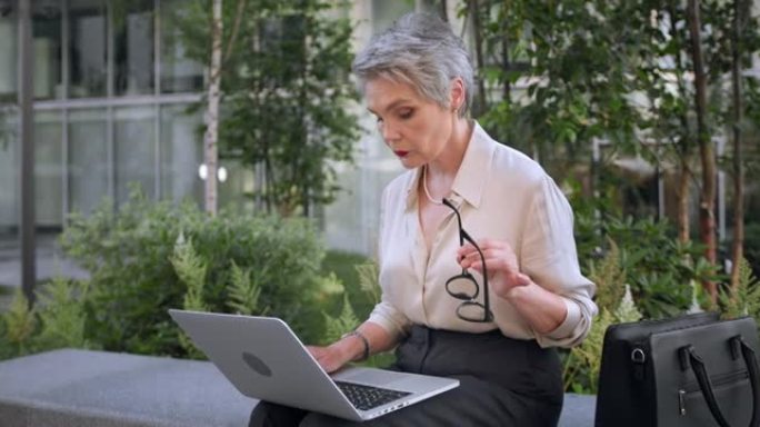 一位才华横溢的白发女作家正在笔记本电脑上创作一本新书，手里转动着眼镜，坐在商务中心前