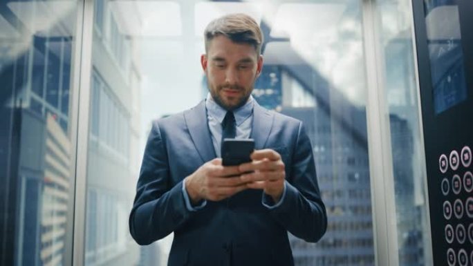 成功的商人在现代商务中心乘坐玻璃电梯到办公室。英俊的快乐男人在使用智能手机时微笑，写短信，查看社交媒