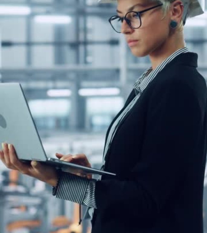 一位戴着白色安全帽的女工程师的垂直肖像，使用笔记本电脑，在汽车装配厂的办公室外面望去。在技术设施中从