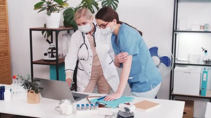 团队合作理念。穿着蓝色磨砂膏的年轻护士咨询，看着笔记本电脑，穿着白大褂的白人高级医生。