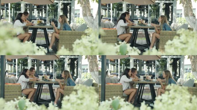 年轻女士朋友坐在户外咖啡馆的餐桌旁聊天和使用智能手机的侧视图