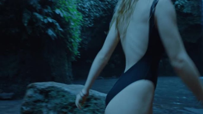 美丽的女人晚上在瀑布边脱衣服，独自在雨林游泳池里游泳，穿着泳衣享受平静的夜晚
