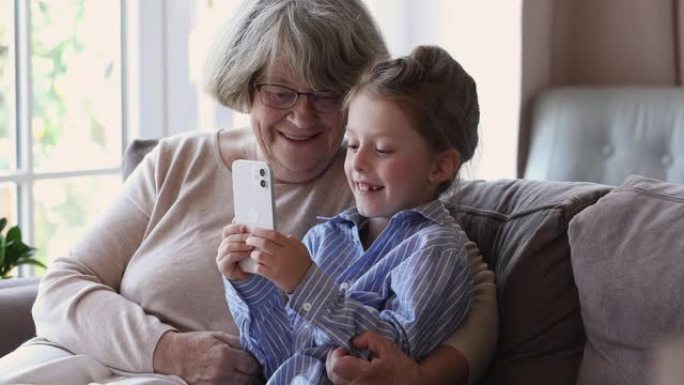 年长的奶奶她的曾孙女用智能手机坐在沙发上