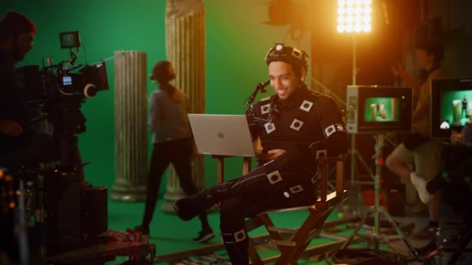 英俊的微笑演员穿着运动捕捉服和头部装备午休，坐在椅子上，使用笔记本电脑。工作室高预算电影。论电影制片