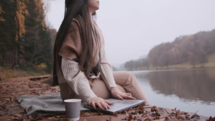 年轻女子坐在湖边的木板路上关闭笔记本电脑盖子