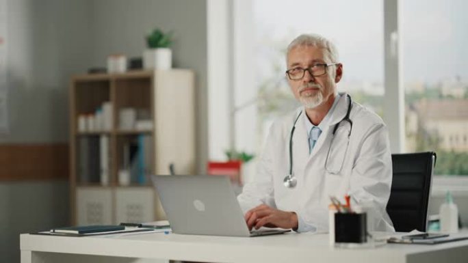 一位经验丰富的中年男医生穿着白大褂在他的办公室用笔记本电脑工作的肖像。资深医疗保健专业人员看着相机，