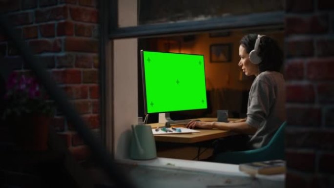 女性设计师使用带有绿屏色度键监视器的台式计算机。远程访问管理器在耳机中听音乐或播客，自由职业者在晚上