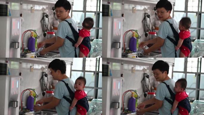 亚洲中国母亲带着她的婴儿在吊带和婴儿背带做家务在厨房里洗碗和杯子，同时和婴儿说话