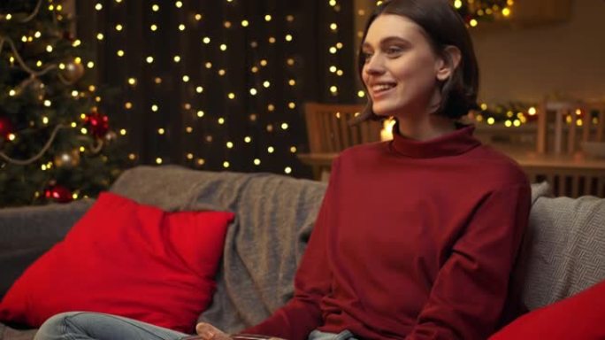 一位穿着红色毛衣的年轻棕色头发的迷人女人正在看电视上的喜剧节目，开玩笑。她坐在装饰精美的圣诞房间的沙