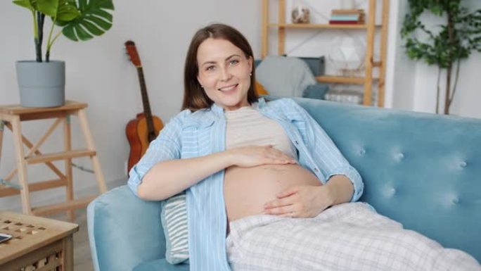 快乐孕妇躺在沙发上抚摸肚子微笑看着相机的肖像