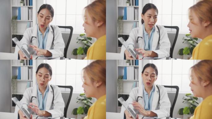年轻的亚洲女性医生在白色医疗制服使用平板电脑正在传递伟大的新闻谈话讨论结果或症状与女孩患者坐在办公桌