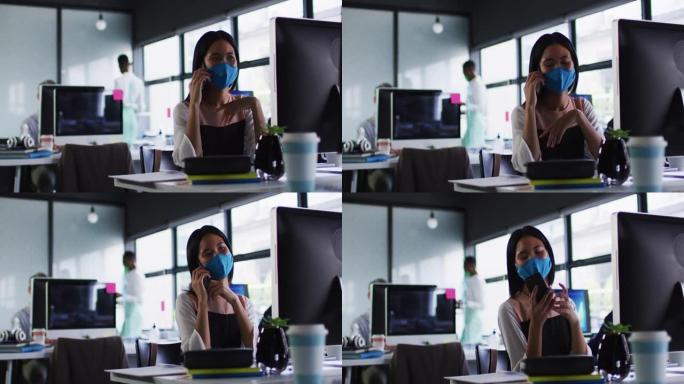 坐在现代办公室的桌子上戴着口罩的亚洲妇女在智能手机上聊天