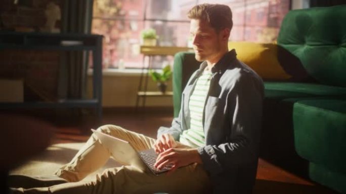 一个迷人的年轻成年男性坐在地板上，在阳光明媚的阁楼客厅使用笔记本电脑的肖像。成功的人在家在光明公寓工