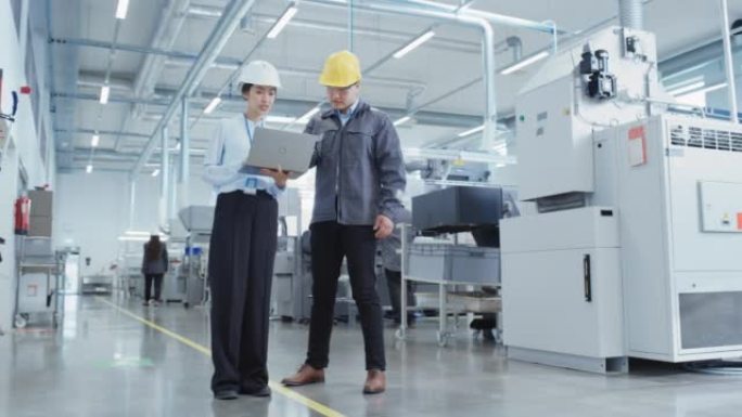 两名年轻的亚洲重工业工程师戴着安全帽，站在笔记本电脑上，在工厂里聊天。两名制造业员工在生产设施工作。