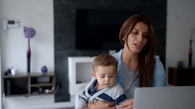 年轻的母亲在家工作，在商务会议上使用笔记本电脑聊天时，将婴儿抱在膝盖上