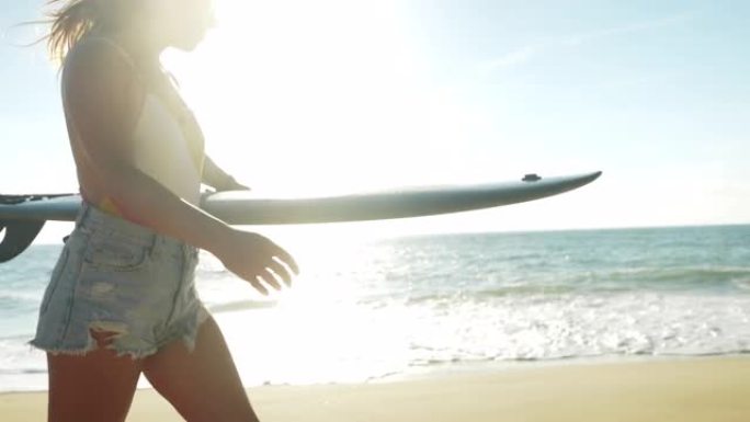 长发的年轻女子，日出时在海滩上散步，手臂下背着冲浪板