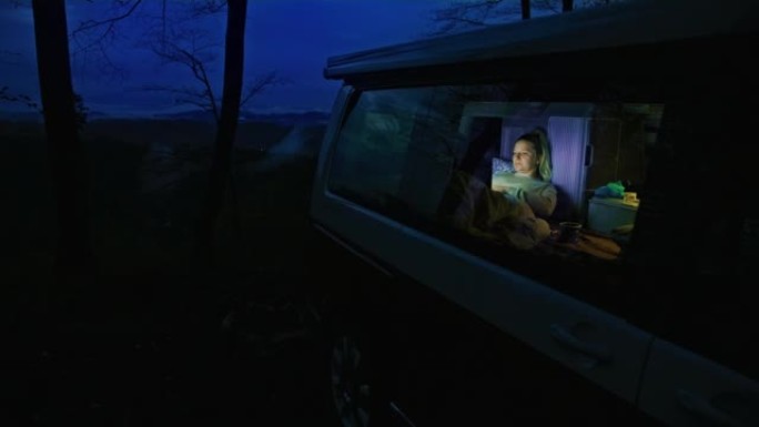 金发碧眼的女人放下杯子，晚上躺在面包车里时停止在笔记本电脑上工作，从户外透过窗户观看