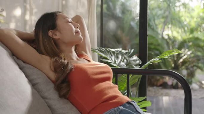 中等镜头: 年轻迷人的宁静亚洲妇女手握在脑后闭上眼睛放松呼吸坐在客厅、阳台或露台上，绿色植物热带休闲