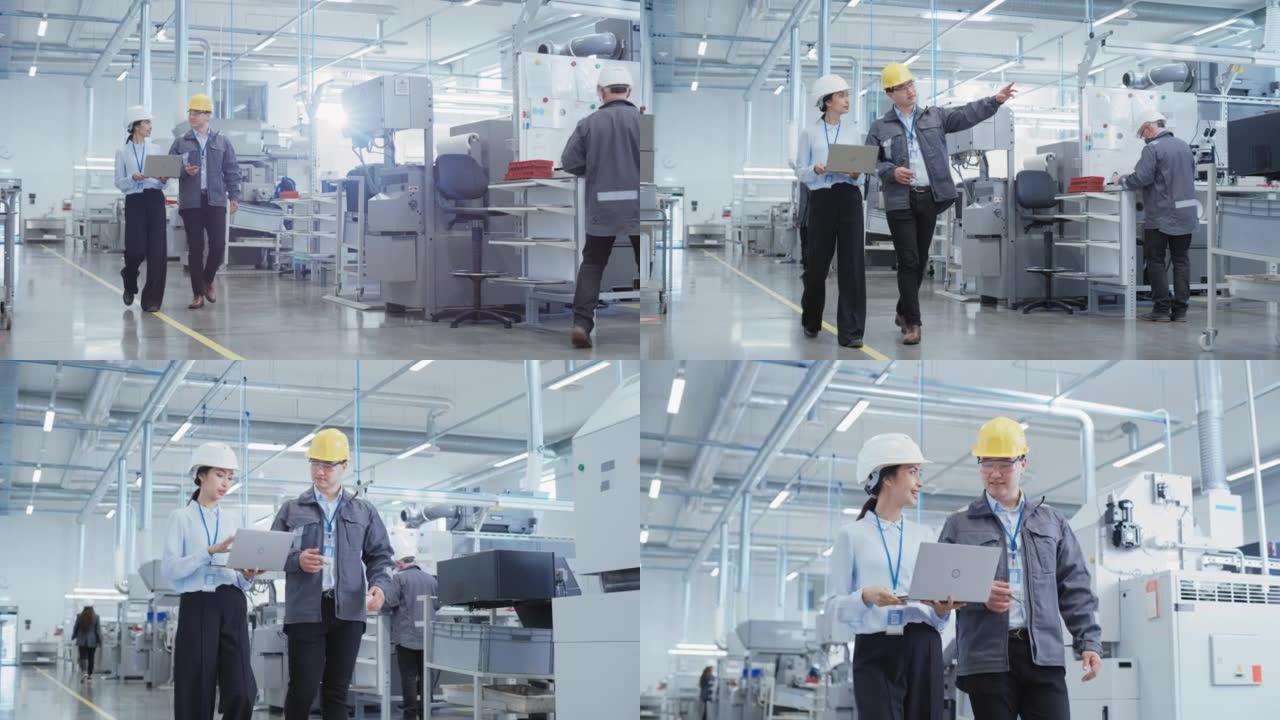 两名年轻的亚洲重工业工程师戴着安全帽，带着笔记本电脑在工厂里散步。两名制造业员工在研发机构工作。