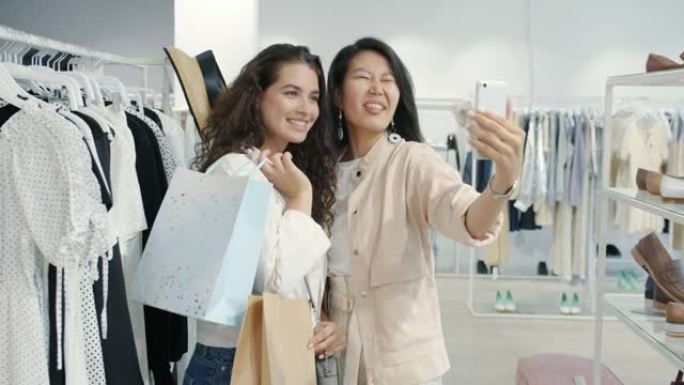 亚洲和高加索女性朋友在服装店用智能手机相机拿着包自拍