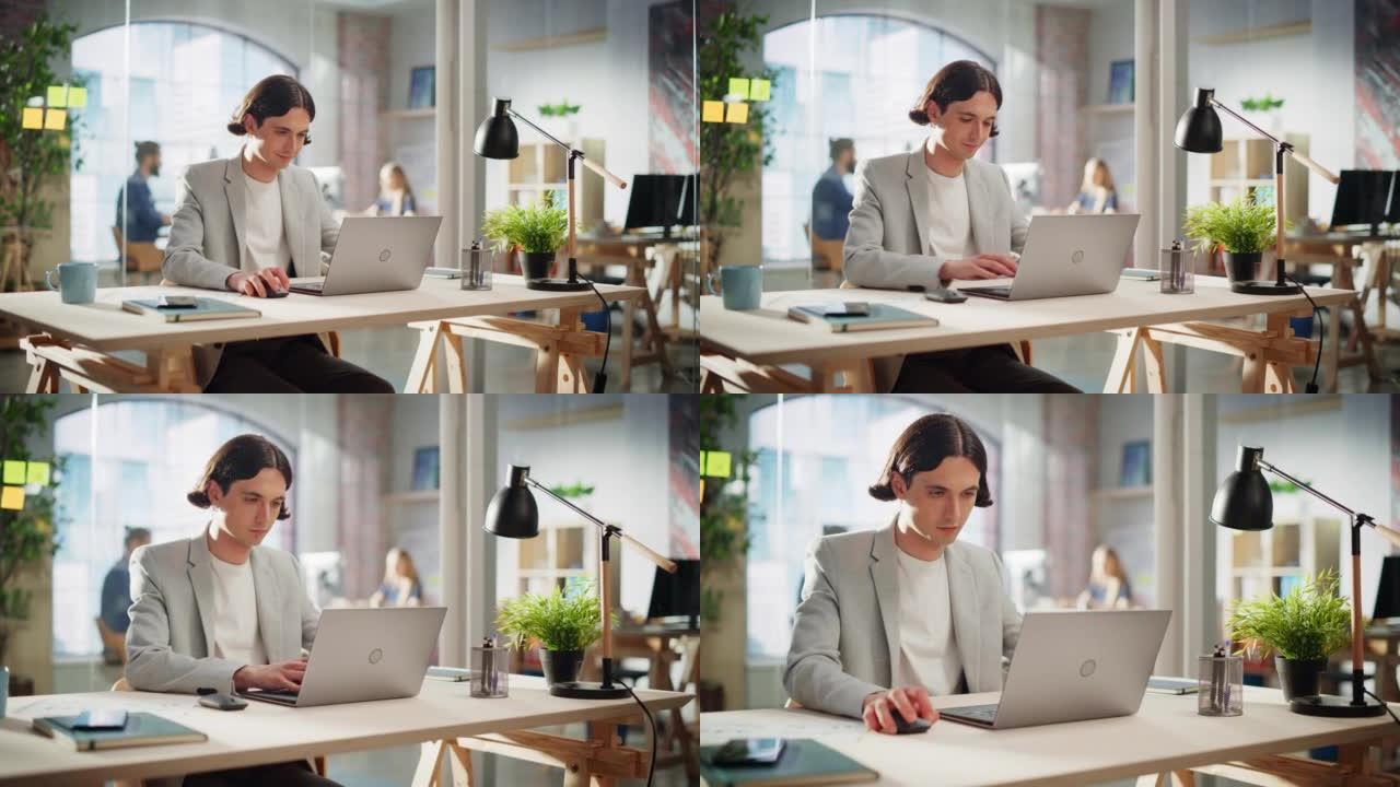快乐的长发经理坐在创意办公室的办公桌前。年轻的时尚男人在营销机构使用笔记本电脑。在后台工作的同事。
