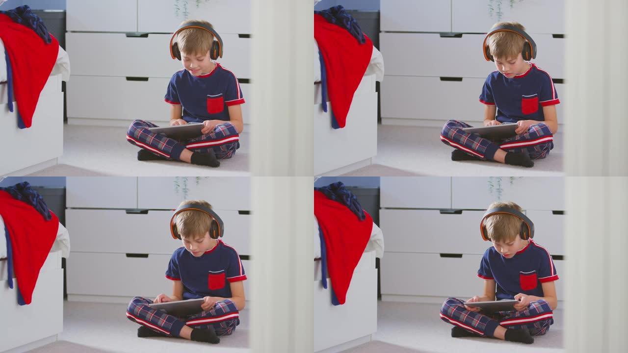男孩在卧室里戴着无线耳机在坐在地板上的数字平板电脑上播放