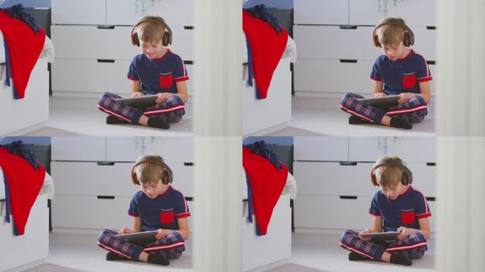 男孩在卧室里戴着无线耳机在坐在地板上的数字平板电脑上播放