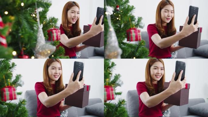 年轻的亚洲女性使用智能手机视频通话与夫妇用圣诞礼物盒交谈，圣诞树装饰着家里客厅的装饰品。