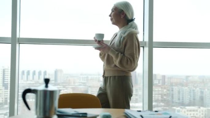 在家庭办公室的全景窗口中，沉思的女士喝咖啡并观看城市景观的肖像