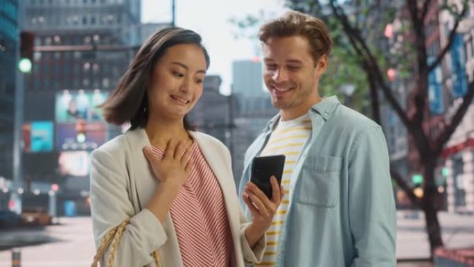 年轻时尚的多民族夫妇站在大城市的街道上。迷人的日本女性向英俊的高加索男性展示智能手机。不同的朋友喜欢