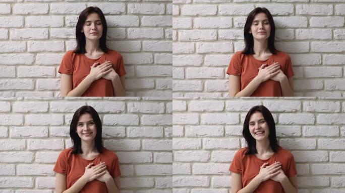 女人将折叠的手掌放在心脏上，在墙壁背景上摆姿势
