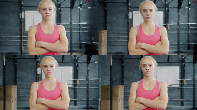 穿着运动服的漂亮金发女孩的肖像站在健身房里，脸严肃，看着相机
