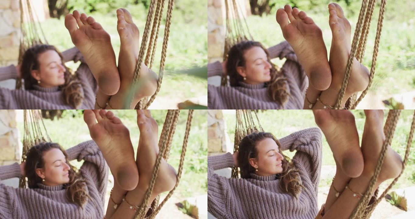 微笑的白人妇女赤脚躺在吊床上，在阳光明媚的花园里闭着眼睛放松