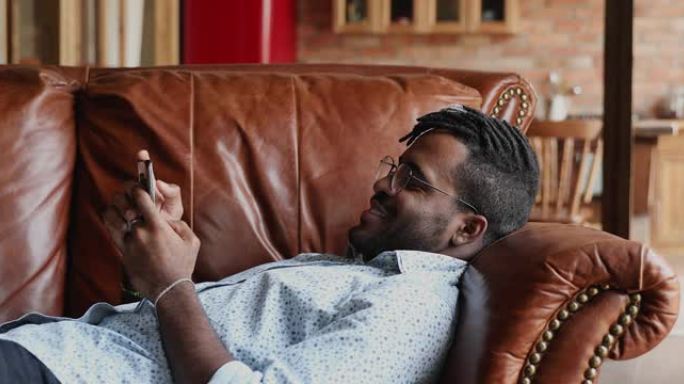 轻松的非洲男子躺在家里的沙发上，用智能手机