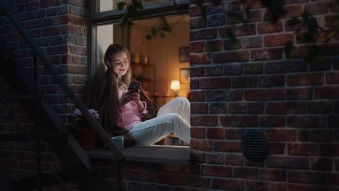 快乐美丽的白人女人晚上坐在窗台上使用智能手机。她正在浏览互联网并在社交网络上查看视频，并从中获得乐趣