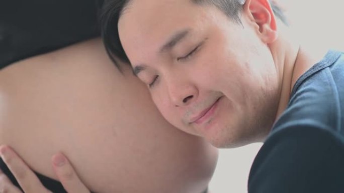 亚洲华裔男子听着，靠在妻子的腹部，表达对他新生的爱和期待