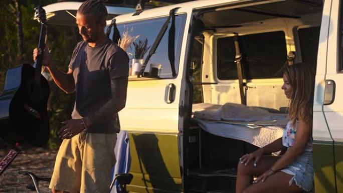 夫妇坐在白色露营面包车的侧面，男人弹吉他，享受日落