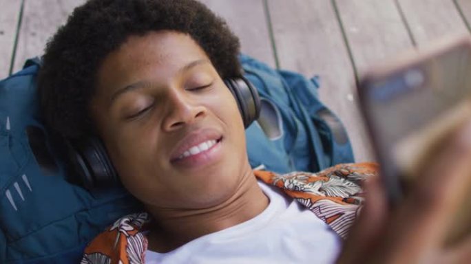 城市里微笑的非洲裔美国人，戴着耳机躺在甲板上，在公园里使用智能手机