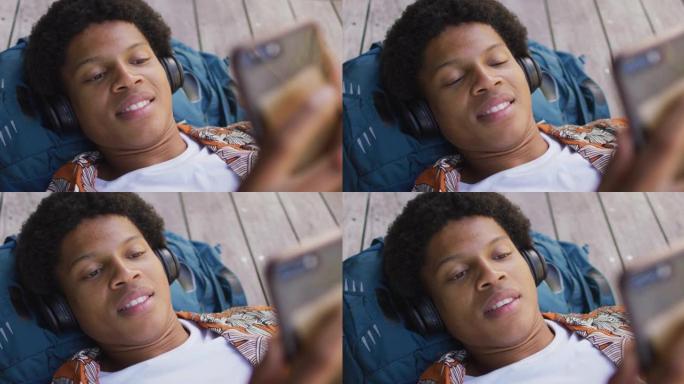 城市里微笑的非洲裔美国人，戴着耳机躺在甲板上，在公园里使用智能手机