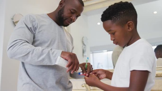 非裔美国爸爸在家里的浴室里把牙膏涂在儿子的刷子上