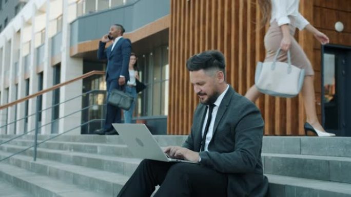 雄心勃勃的中年企业家使用笔记本电脑坐在现代城市办公楼附近的楼梯上