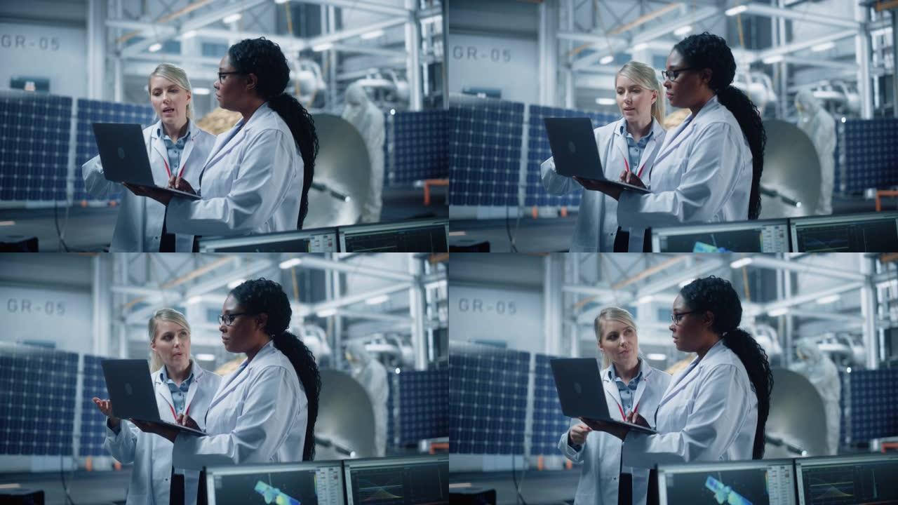 两名女工程师交谈，使用计算机在航空航天卫星制造工厂工作。由多民族科学家组成的多元化团队为太空探索计划