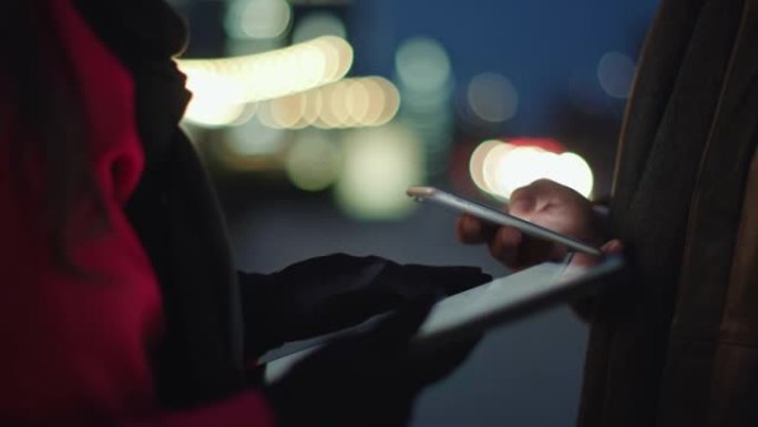 女士女士使用平板电脑，而晚上在城市旁边的男人使用智能手机