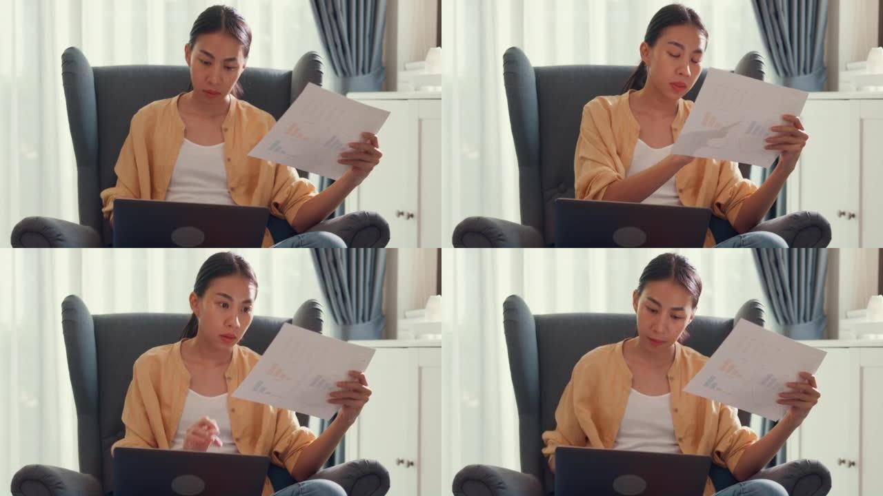 年轻迷人的亚洲女学生或大学生坐在沙发椅上，用电脑笔记本电脑检查文件中的数据图表在家里的客厅报告工作。
