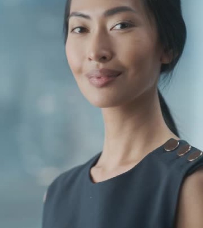 一位亚洲女商人的垂直肖像，穿着时髦的黑色连衣裙，使用笔记本电脑，在城市办公室的窗户旁边摆姿势。美丽的