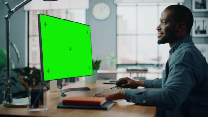 英俊的黑人非洲裔美国专家在家用客厅的台式电脑上工作，绿屏模拟显示。自由职业者在社交网络上通过互联网与