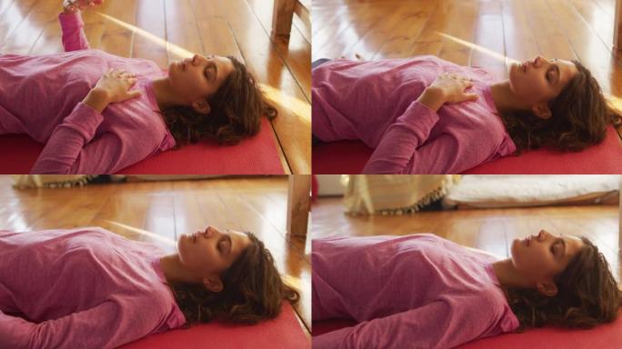 放松的混血女人练习瑜伽，躺在垫子上闭着眼睛在阳光明媚的小屋卧室